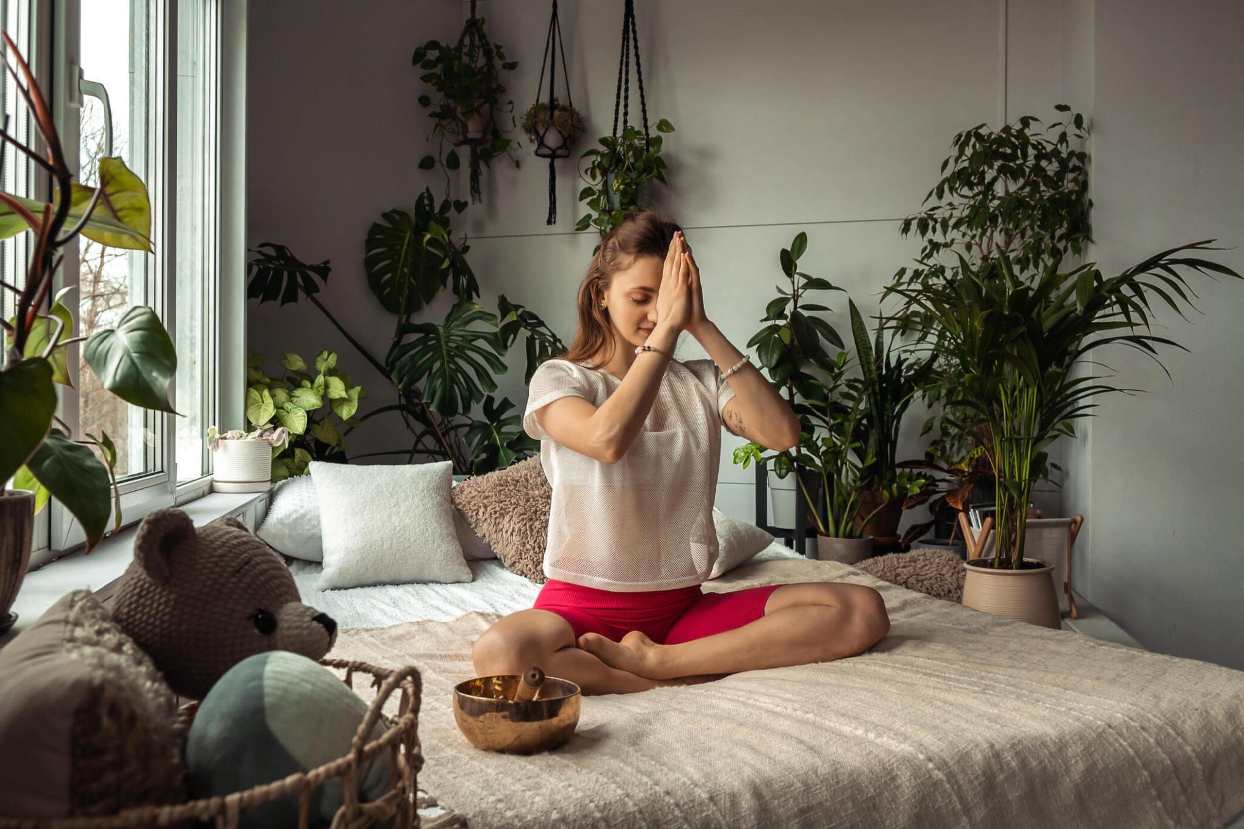 sound therapy woman sitting cross legged hands in prayer with singing bowl on bed surrounded by plants