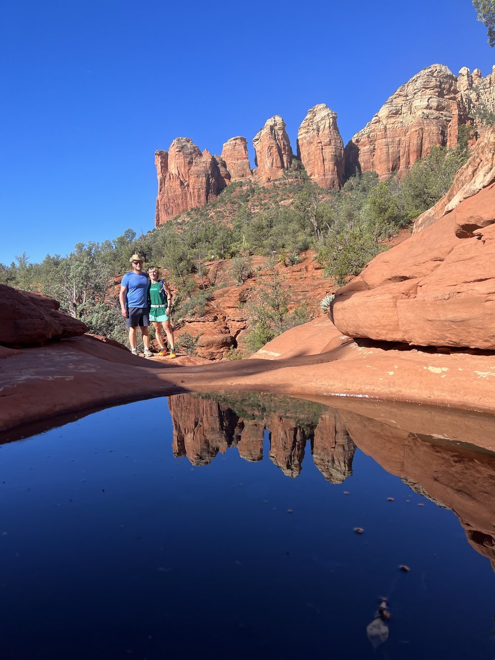 two people on a hiking trail in Sedona Arizona. red rock formations are seen in the background with water in the foreground.
