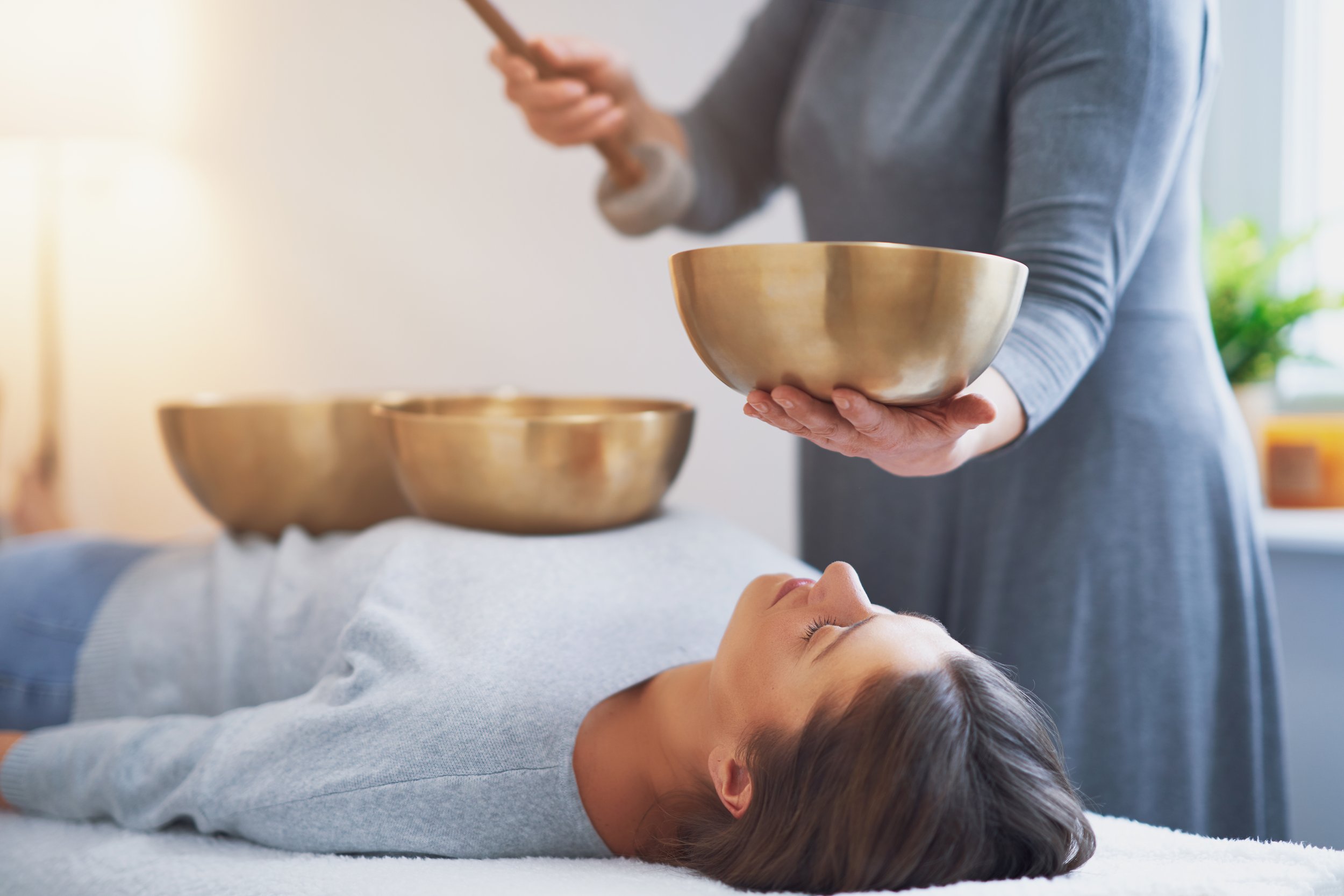 woman receiving sound therapy is resting. sound healing practitioner is scene nearby with singing bowl.