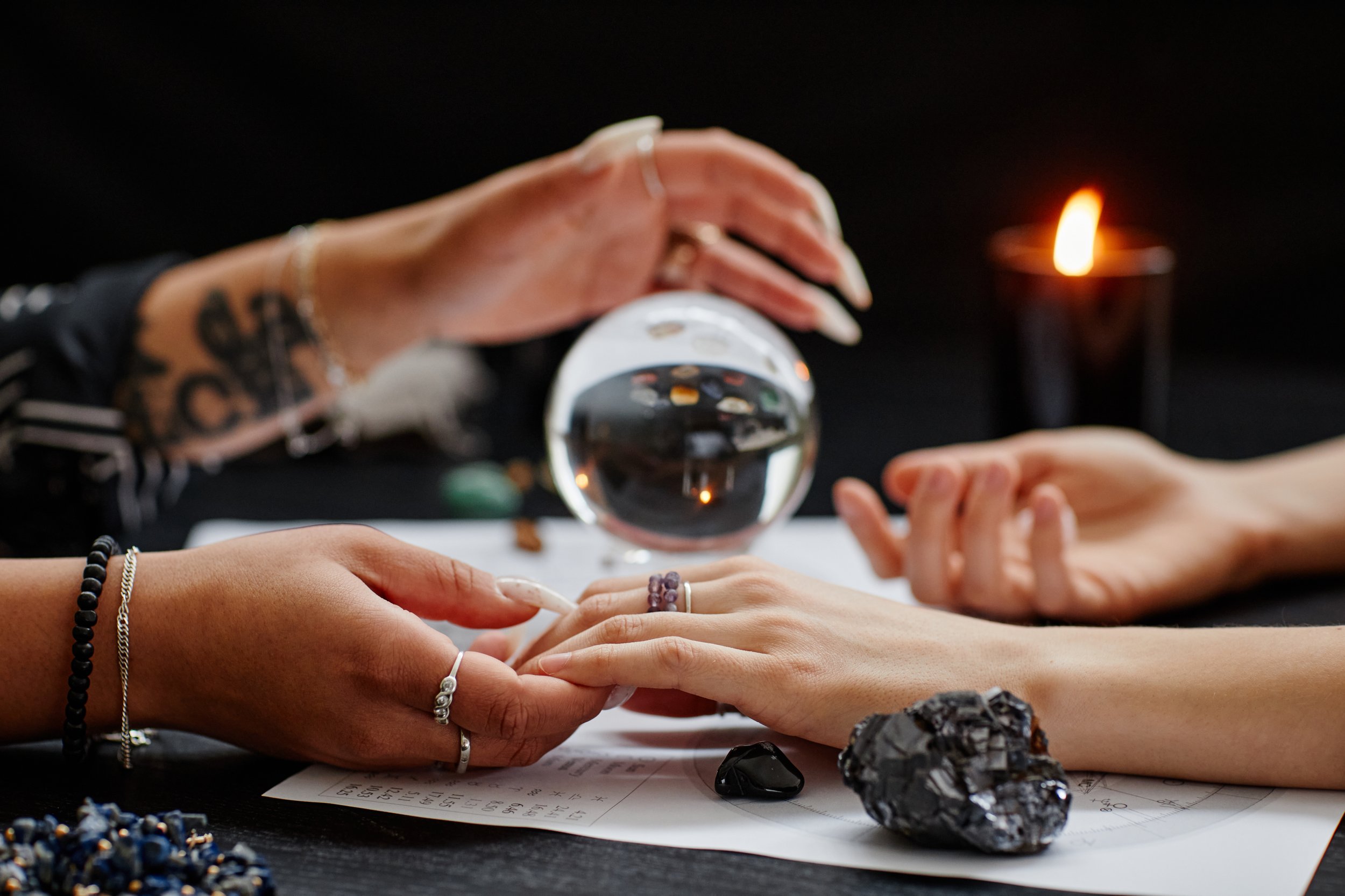 psychic reader's hand is holding fingertips of a woman with both of their hands placed on a table. crystal ball in background.