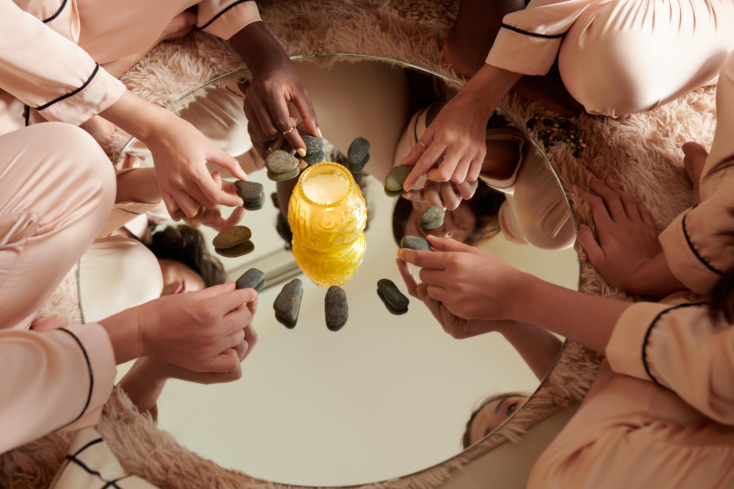 stones are placed in a circle around a central candle which situs on top of mirror. women's hands are touching different stones.